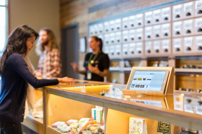 Customer browsing products at a modern cannabis dispensary counter with a digital menu display in the foreground and staff assisting shoppers in the background.
